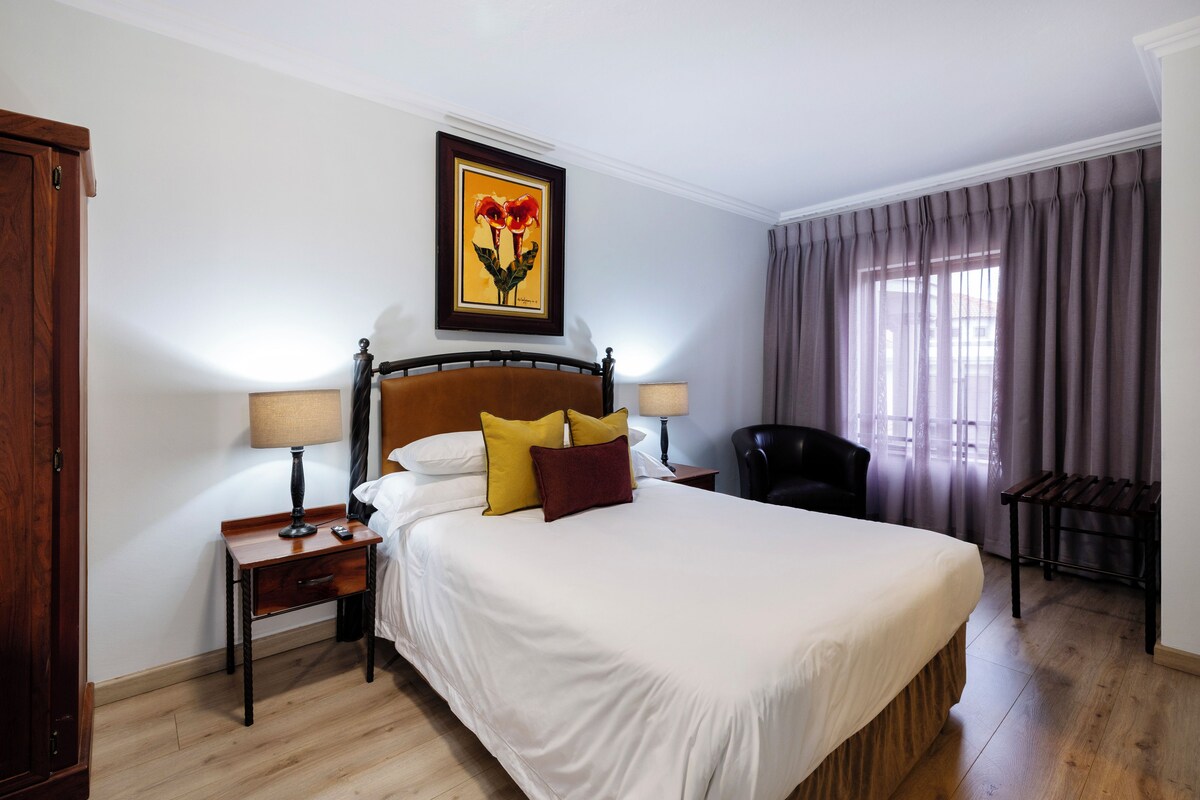 Habitación de hotel con una cama grande, dos mesitas de noche, una silla, una ventana con cortinas y un cuadro en la pared.