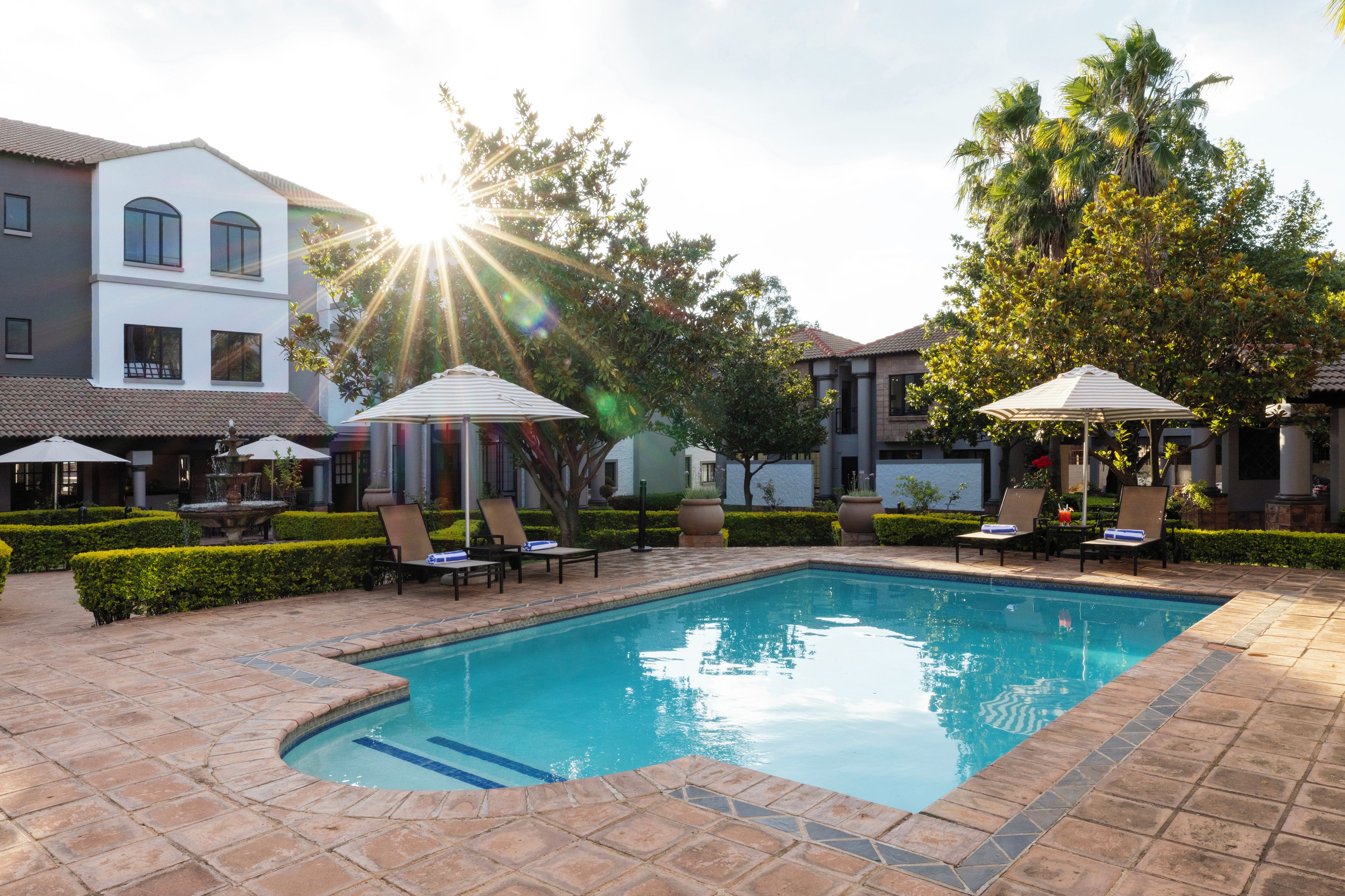 A swimming pool with lounge chairs and umbrellas, surrounded by buildings and trees.