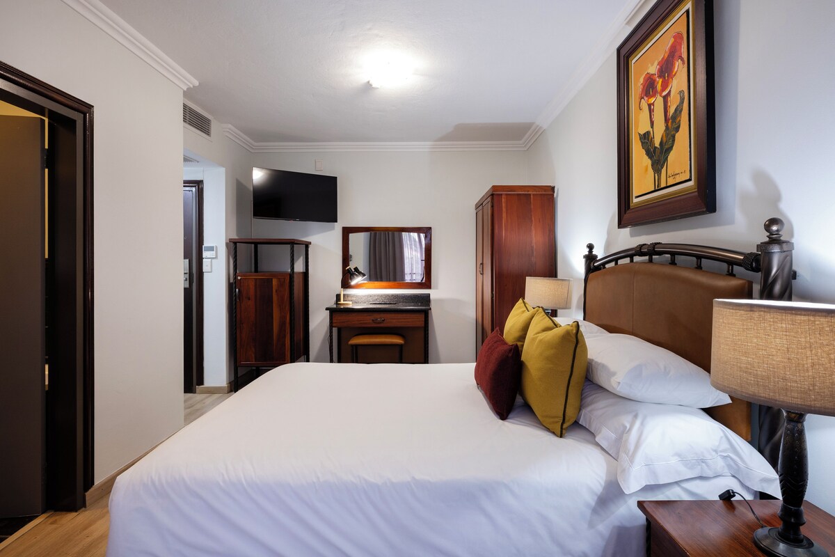 Una habitación de hotel con cama, escritorio, silla, televisor, armario y un cuadro en la pared.