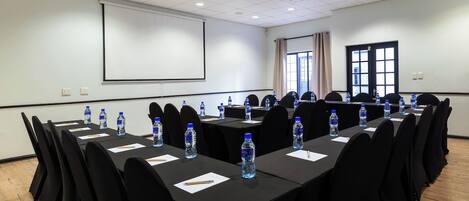 Un salón de conferencias con mesas largas, sillas negras, botellas de agua y una pantalla de proyector.