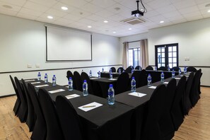 Un salón de conferencias con mesas largas, sillas negras, botellas de agua y una pantalla de proyector.