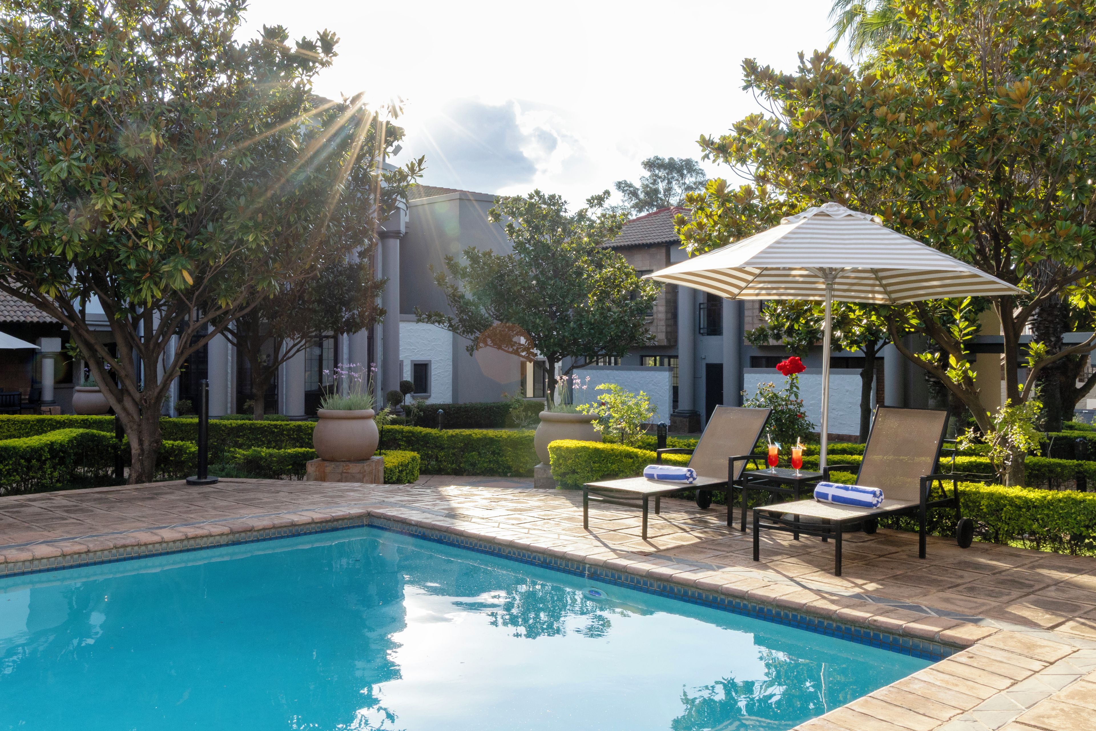 Piscina com espreguiçadeiras e guarda-sol, rodeada por vegetação e edifícios.