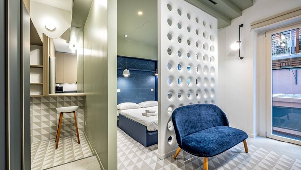 Ein modernes Schlafzimmer mit einem geometrisch gemusterten Boden, einem blauen Sessel, einem Bett mit weißen Bettwäsche und einer Wand mit runden Aussparungen.