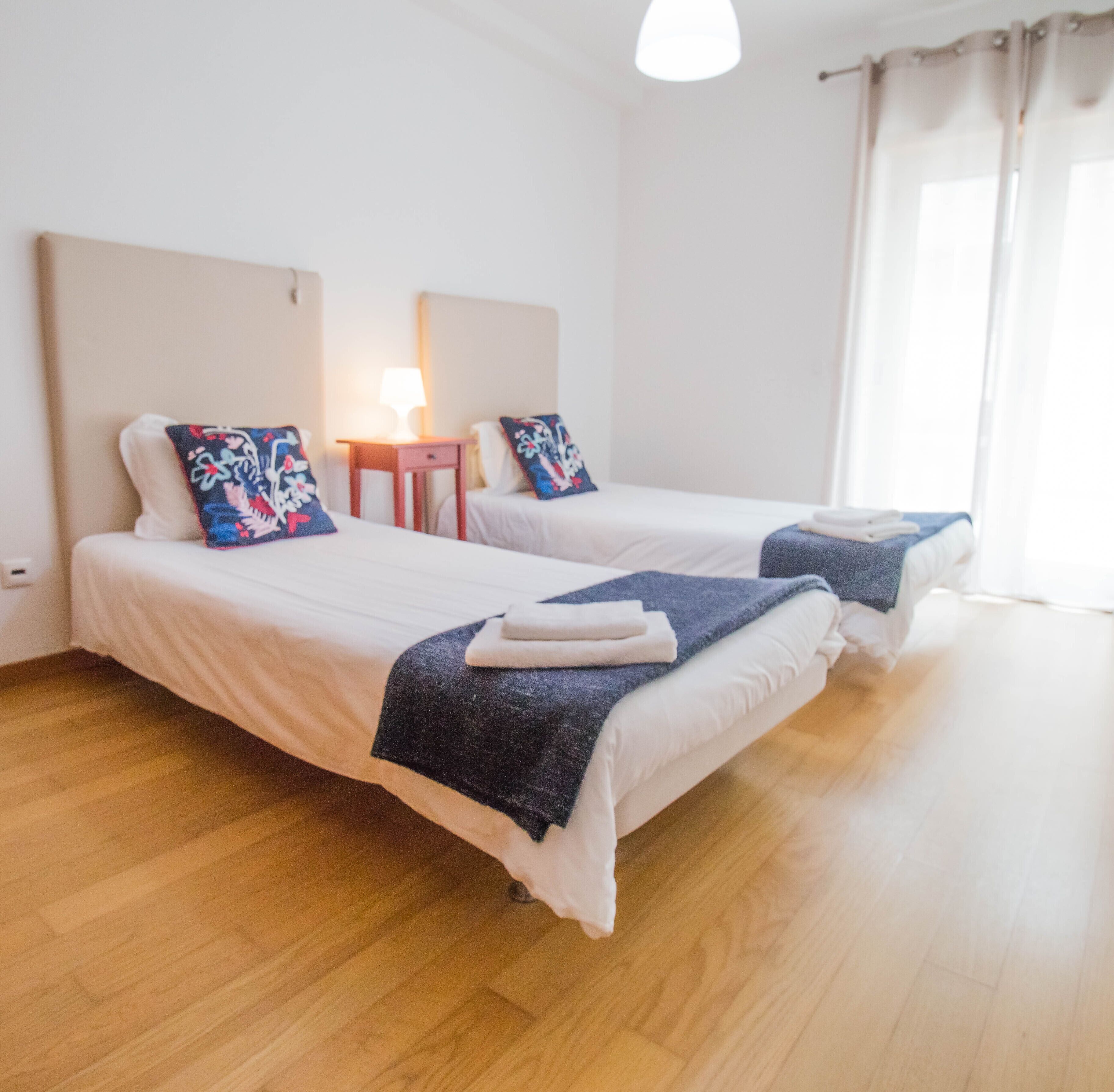 Hotellihuone, jossa on kaksi erillistä sänkyä, pieni yöpöytä lampun kanssa ja ikkuna, jossa on verhot.
