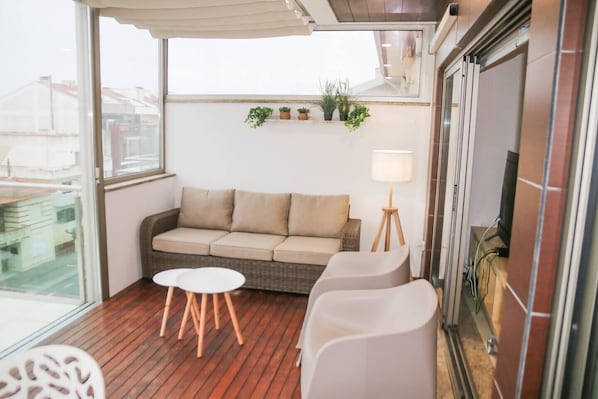 Un balcón con un sofá, dos sillones, una mesita y una lámpara. Hay plantas en macetas en la pared.