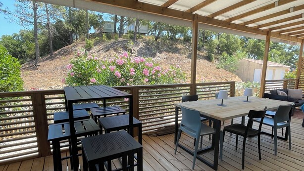 Um pátio externo com mesas e cadeiras de madeira, uma pérgola acima e vista para um morro com vegetação e uma casa ao fundo.