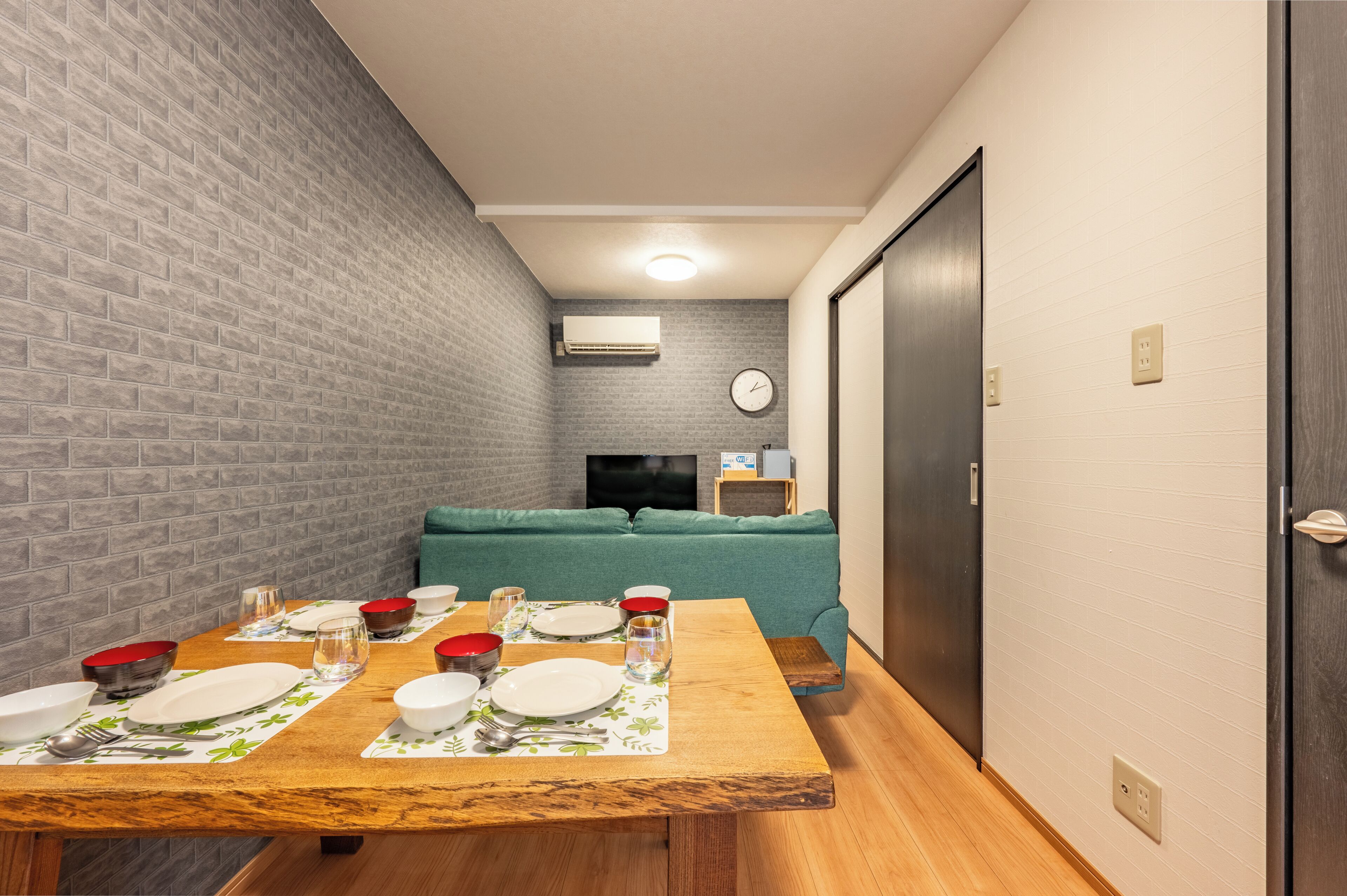 A dining area with a brick wall, a wooden table set with plates and bowls, a green sofa, and a TV in the background.
