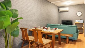 A living room with a brick wall, a green sofa, wooden furniture, and a large plant. - 外観は古い建物ですが内装はフルリノベーション済みです。TV、無料Wi-Fi、ドライヤー、浴室乾燥機、洗濯機完備。トイレにはウォシュレットも完備しております。 (Nakamichi,Higashinari-ku,Osaka-shi)
