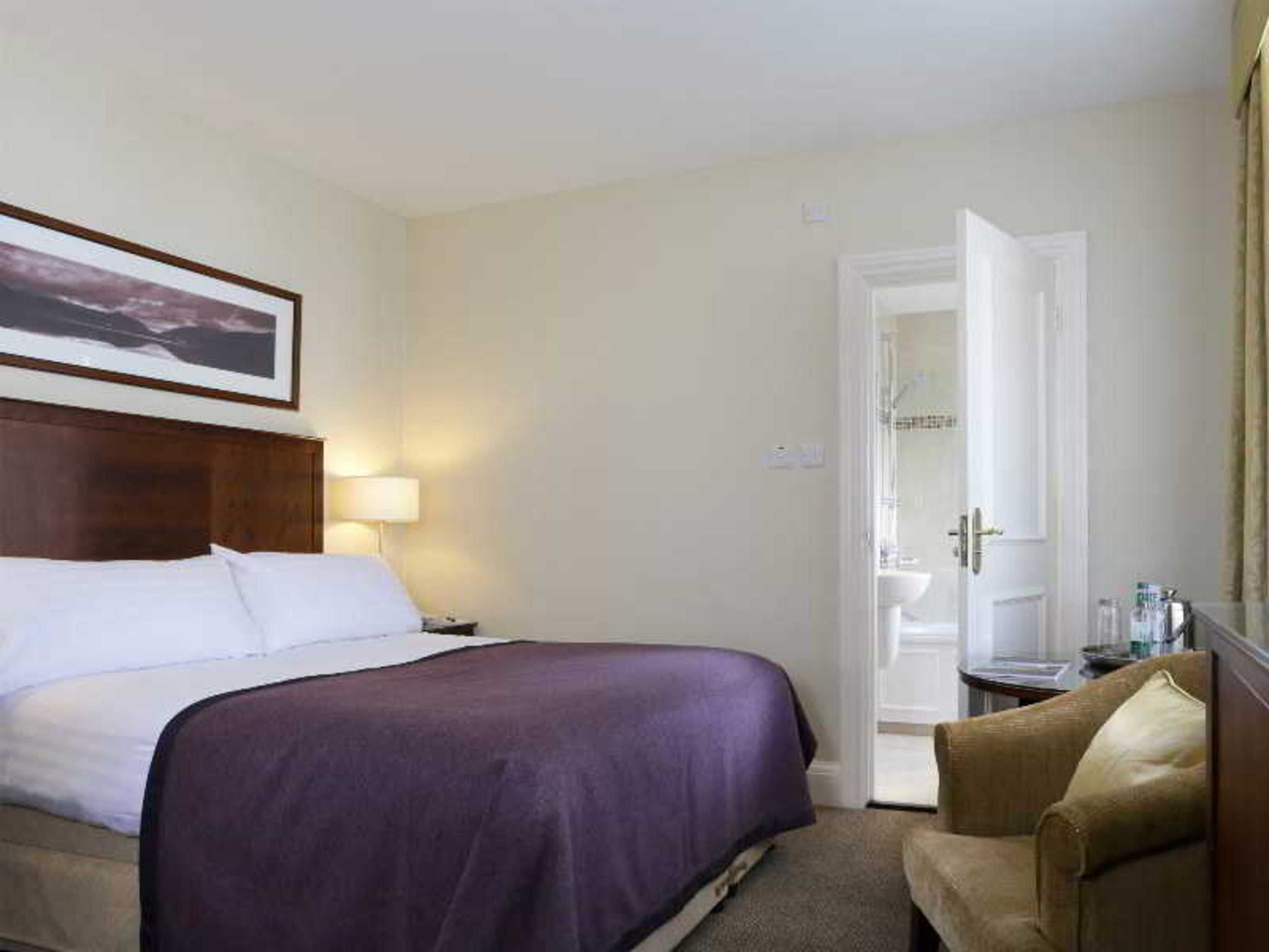 A hotel room with a bed, a chair, a nightstand, a lamp, and a door leading to a bathroom.