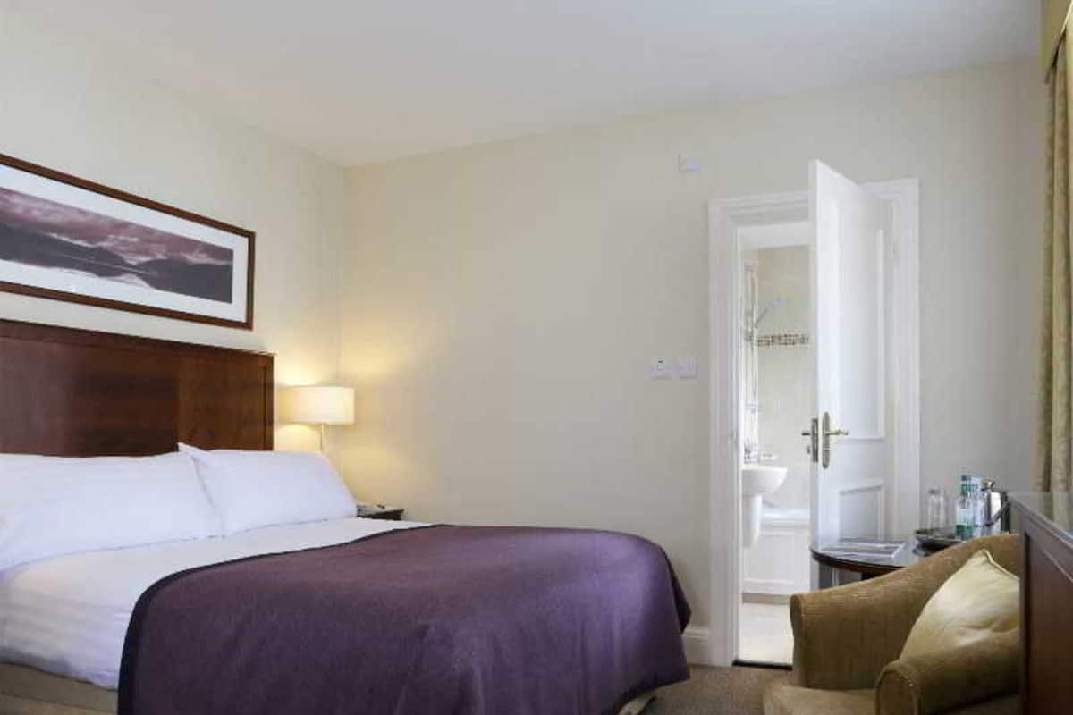Una habitación de hotel con cama, una silla, mesita de noche, lámpara y puerta que da a un baño.