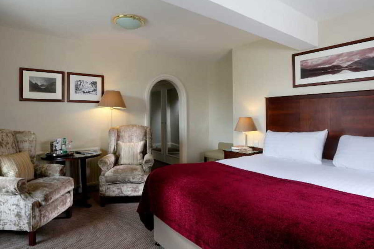 A hotel room with a large bed, two armchairs, a small table, and a nightstand. There are two framed pictures on the wall and a ceiling light.