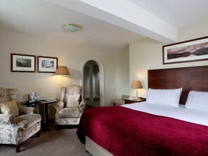 Habitación de hotel con una cama grande, dos sillones, una mesita y una mesita de noche. Hay dos cuadros en la pared y una lámpara en el techo.