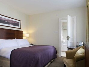 Una habitación de hotel con cama, una silla, mesita de noche, lámpara y puerta que da a un baño.