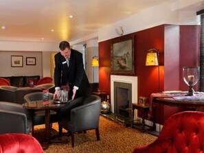 Un vestíbulo de hotel con chimenea, sillas tapizadas de rojo, una mesa redonda con un vaso y un hombre organizando objetos sobre una mesa.