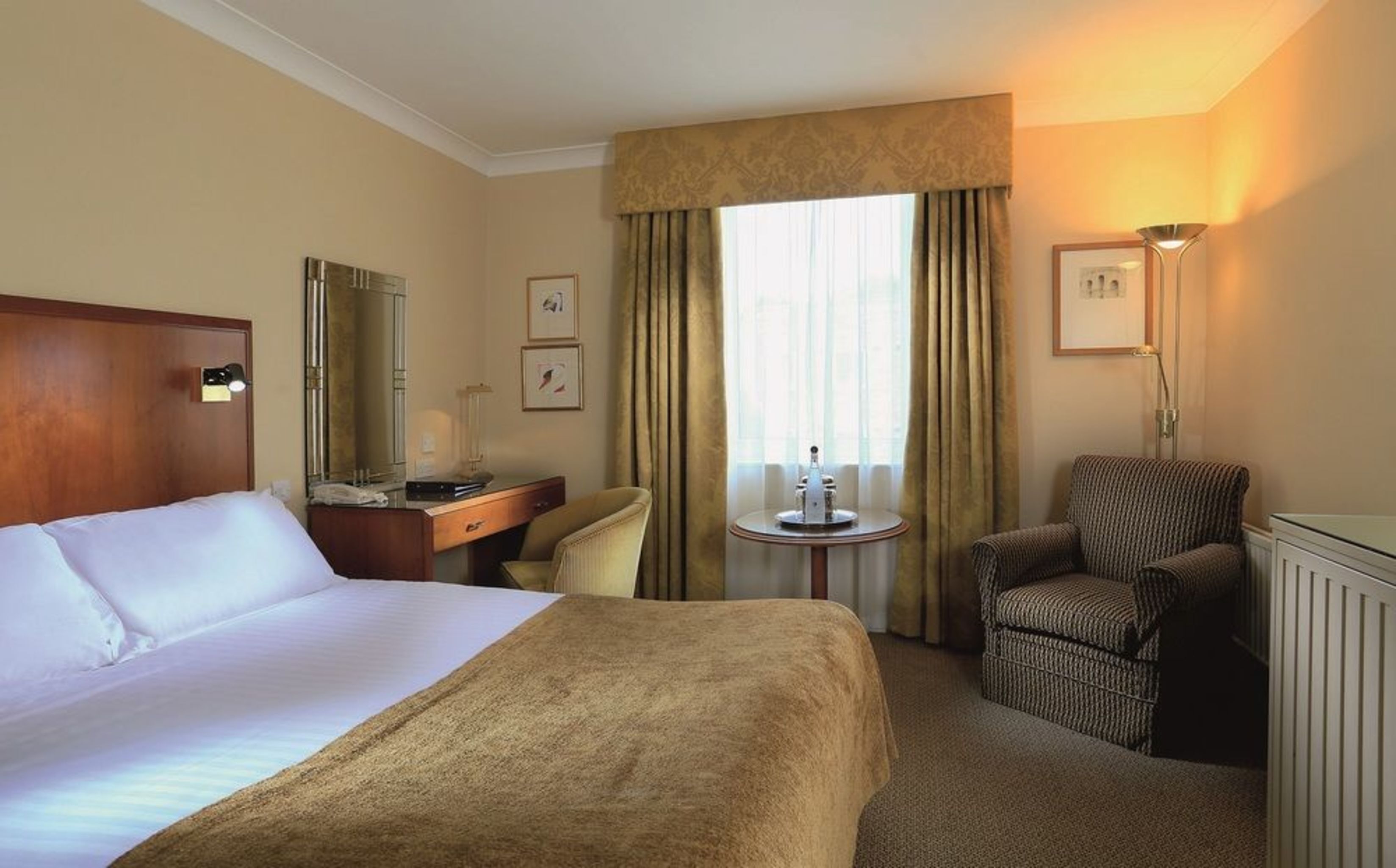 A hotel room with a large bed, a desk, a chair, a small table, and a window with curtains.