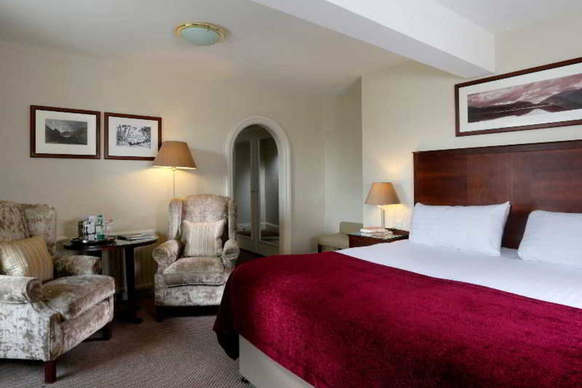 Habitación de hotel con una cama grande, dos sillones, una mesita y una mesita de noche. Hay dos cuadros en la pared y una lámpara en el techo.