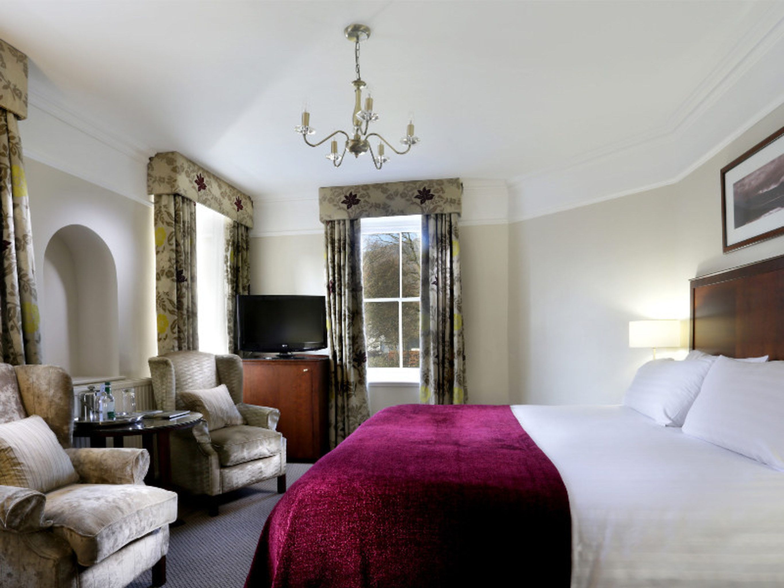 A hotel room with a bed, a television, a desk, and two armchairs.