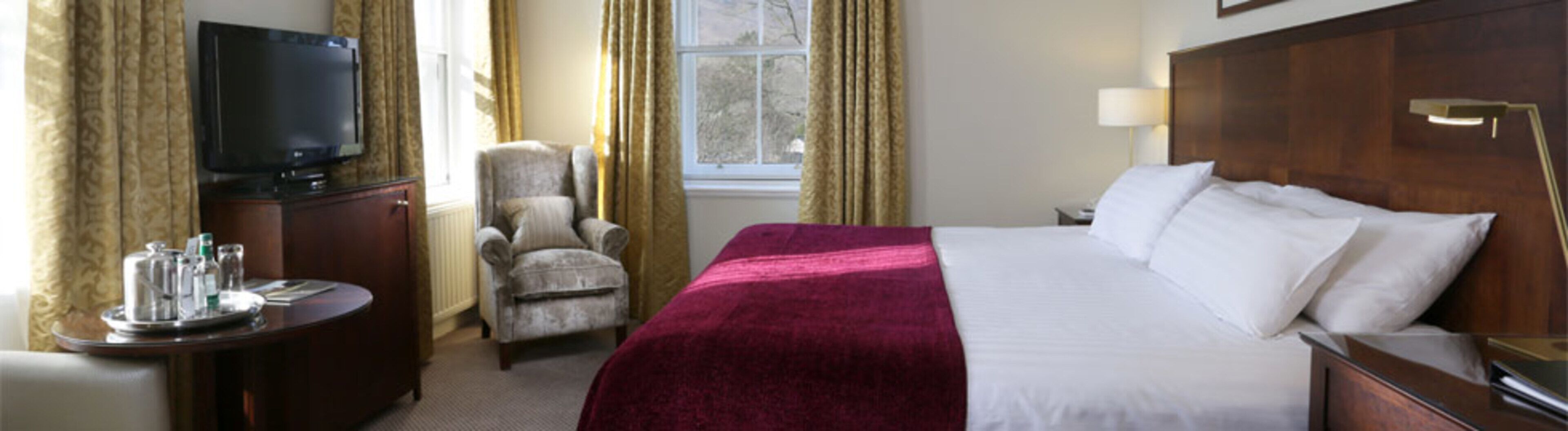 A hotel room with a bed, a TV, a chair, and a desk with a tray of items.