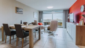 Un espace repas moderne avec une table et des chaises en bois, un coin cuisine agrémenté d’un mur d’accent rouge, et de grandes fenêtres offrant une vue sur l’extérieur.