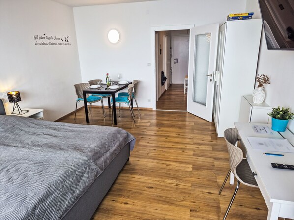 Una habitación moderna con una cama, un escritorio, una silla, una mesa de comedor con sillas y una puerta que da a otra habitación.