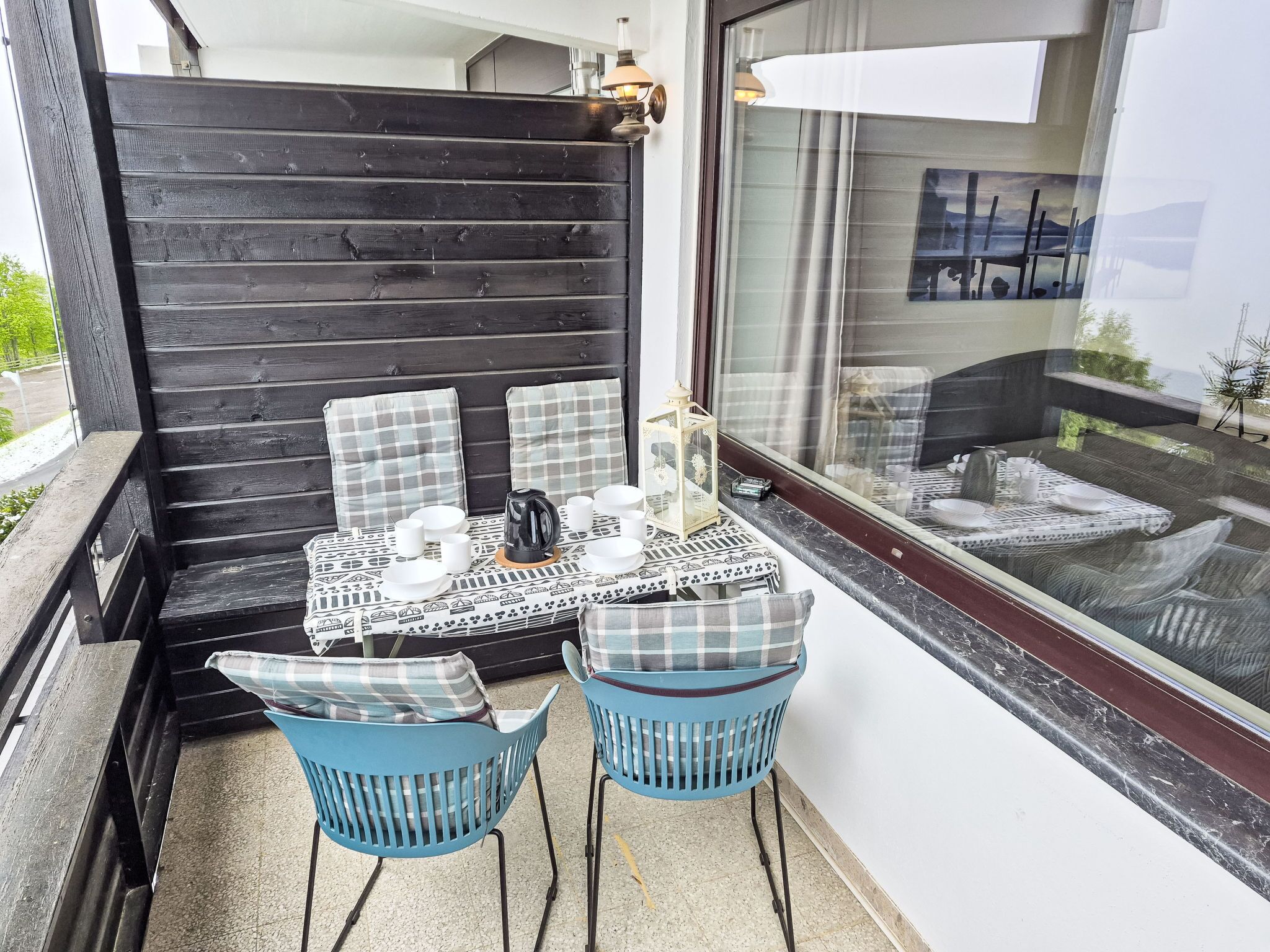 Een terras met een tafel voor twee personen, twee blauwe stoelen, een lantaarn en uitzicht op een tuin door een raam.