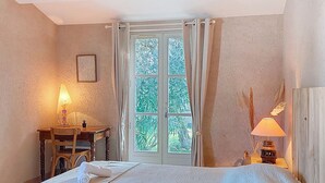 A bedroom with a large bed, a wooden desk with a lamp, a window with curtains, and a view of greenery outside. - Le Château 3 by Interhome (Ile d'Oléron)