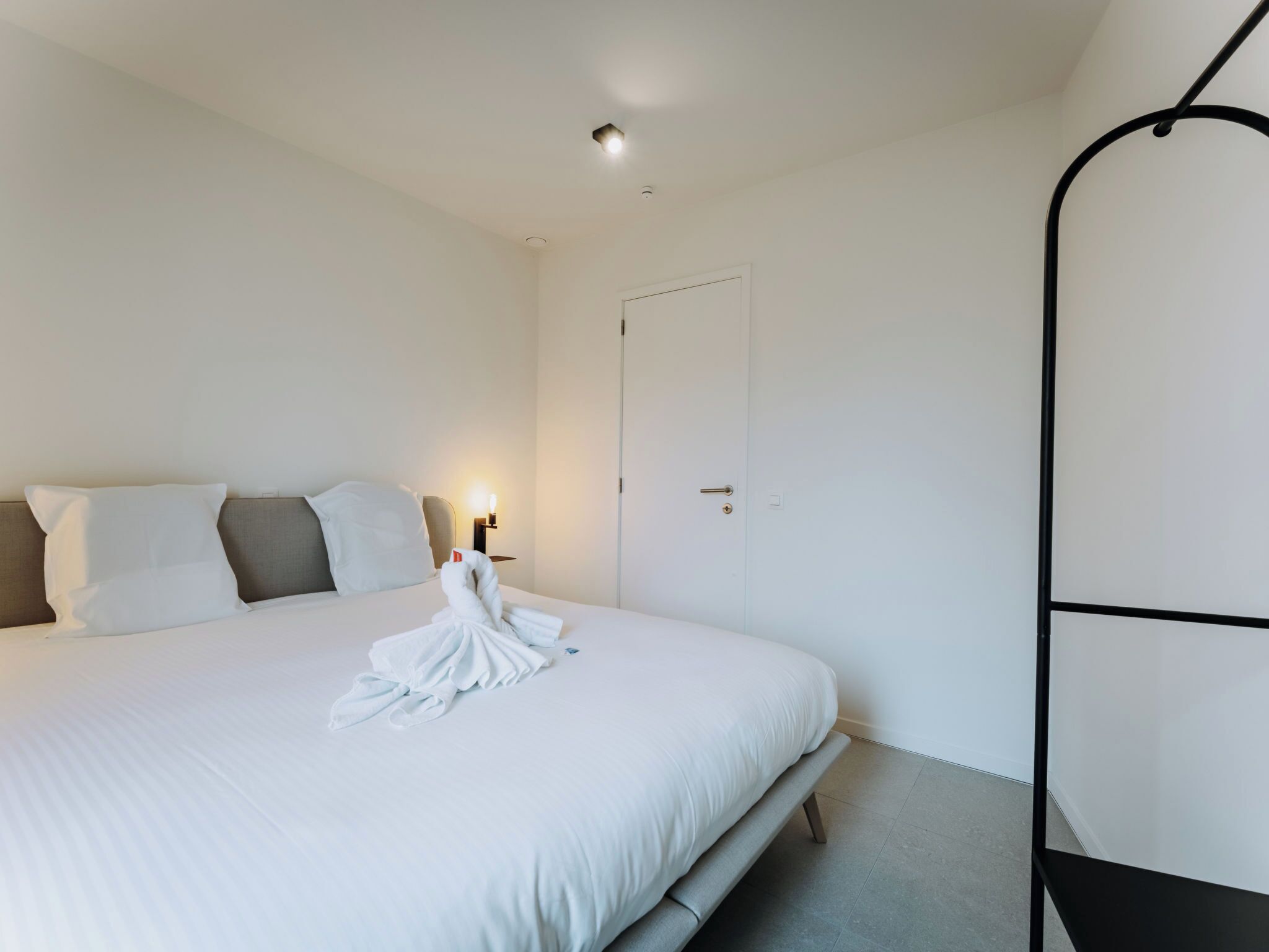 A neatly made bed with white linens and a folded towel, a bedside lamp, and a door with a handle.
