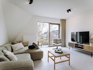 A modern living room with a sofa, armchair, and coffee table. A television is on a stand, and there is a large window with a view of buildings. - West Bay with parking & balcony by Interhome (Westende-Bad)