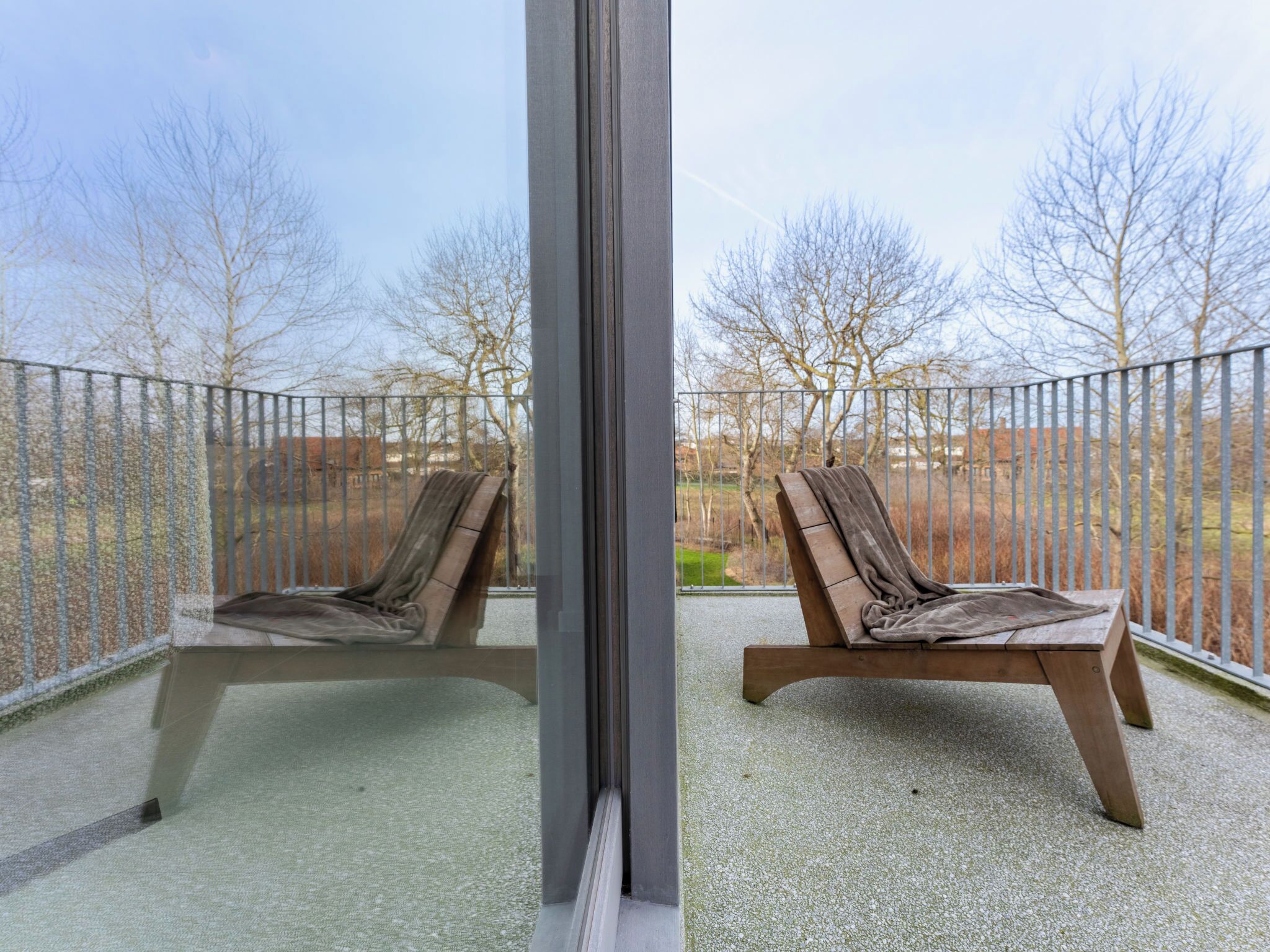 A balcony with two wooden lounge chairs, a metal fence, and a view of trees and buildings in the background.