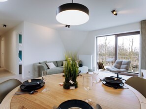 Ein modernes Esszimmer mit einem runden Holztisch, gedeckt für vier Personen, einer grauen Couch und Blick ins Freie durch große Fenster.