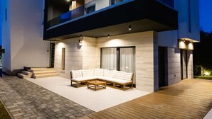 A modern two-story house with a wooden deck, a sectional sofa, and a staircase leading to the entrance.