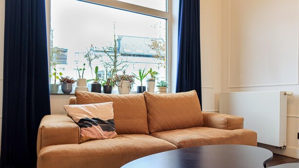 Ein Wohnzimmer mit einem beigen Sofa, einem runden Couchtisch und mehreren Blumentöpfen auf einem Fensterbrett.