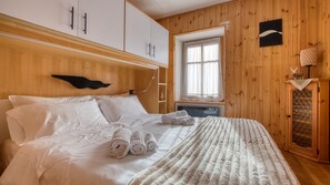 Ett sovrum med en säng, vita sängkläder, ett fönster med gardiner, träpanel på väggarna och ett konstverk som hänger på väggen.