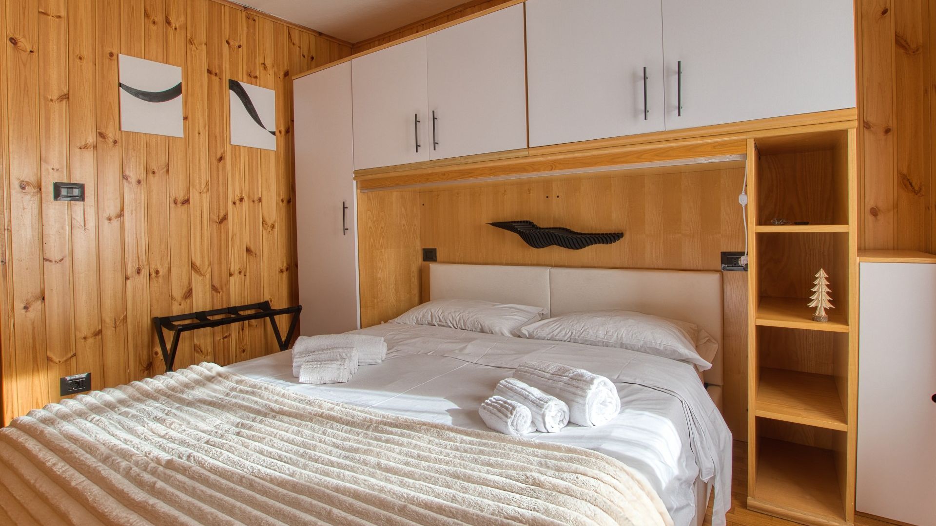 A bedroom with a wooden headboard, a bed with white linens, a nightstand, and a wooden wardrobe with a small Christmas tree decoration.