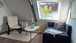 A cozy loft bedroom with a sloped ceiling, a window with a view of trees, a dark sofa with a throw blanket, a chair, and a small table with a vase. - Lütt Stuuv im historischen Flensburger Hafen (Flensburg)