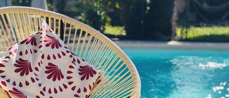 A wicker chair with a patterned cushion by a pool, a metal mug, and a clear view of the pool and palm trees.