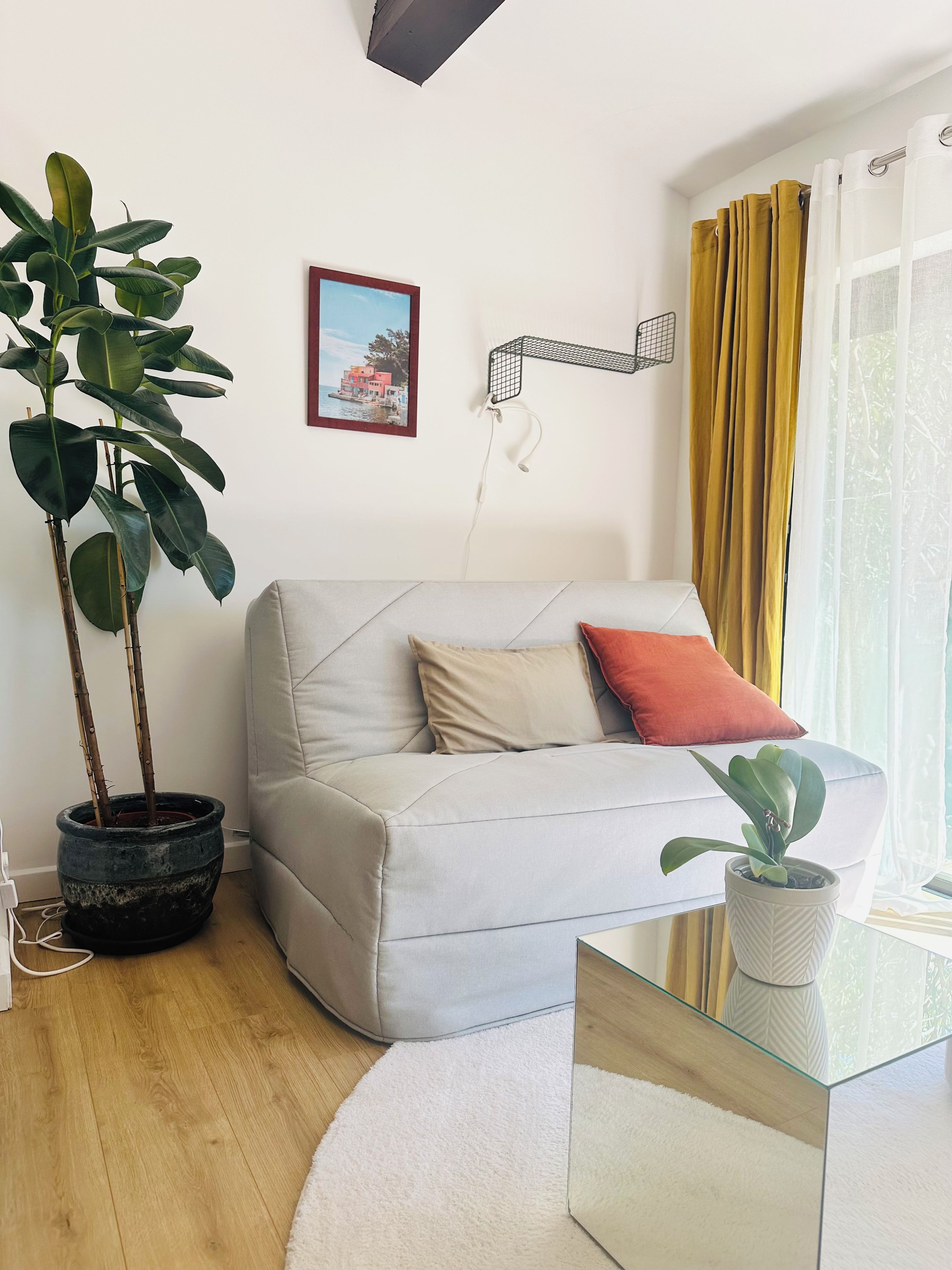 Ein Wohnzimmer mit einer Couch, einem Blumentopf, einem gerahmten Bild und einem Fenster mit Vorhängen.