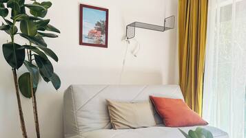 Ein Wohnzimmer mit einer Couch, einem Blumentopf, einem gerahmten Bild und einem Fenster mit Vorhängen.
