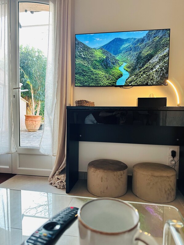 Un salon avec un téléviseur fixé au mur, diffusant un paysage pittoresque, une porte avec des rideaux, une plante en pot et deux poufs.