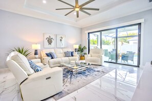 A modern living room with a glass coffee table, white sofas, and a large sliding glass door leading to an outdoor patio. - Bergamo 4BR Pool Home | Hot Tub & Ping Pong (Naples)