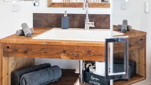 Een badkamer met een houten wastafelmeubel, een grote spiegel en een wastafel. Er liggen handdoeken en staat een zeepdispenser op het meubel.
