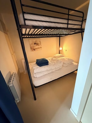 Una cama litera con ropa de cama blanca y una toalla azul encima. Hay un radiador a la izquierda y una lámpara de pared a la derecha.