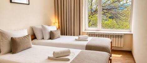 A hotel room with two beds, a window with a view of trees, a radiator, and a framed picture on the wall.