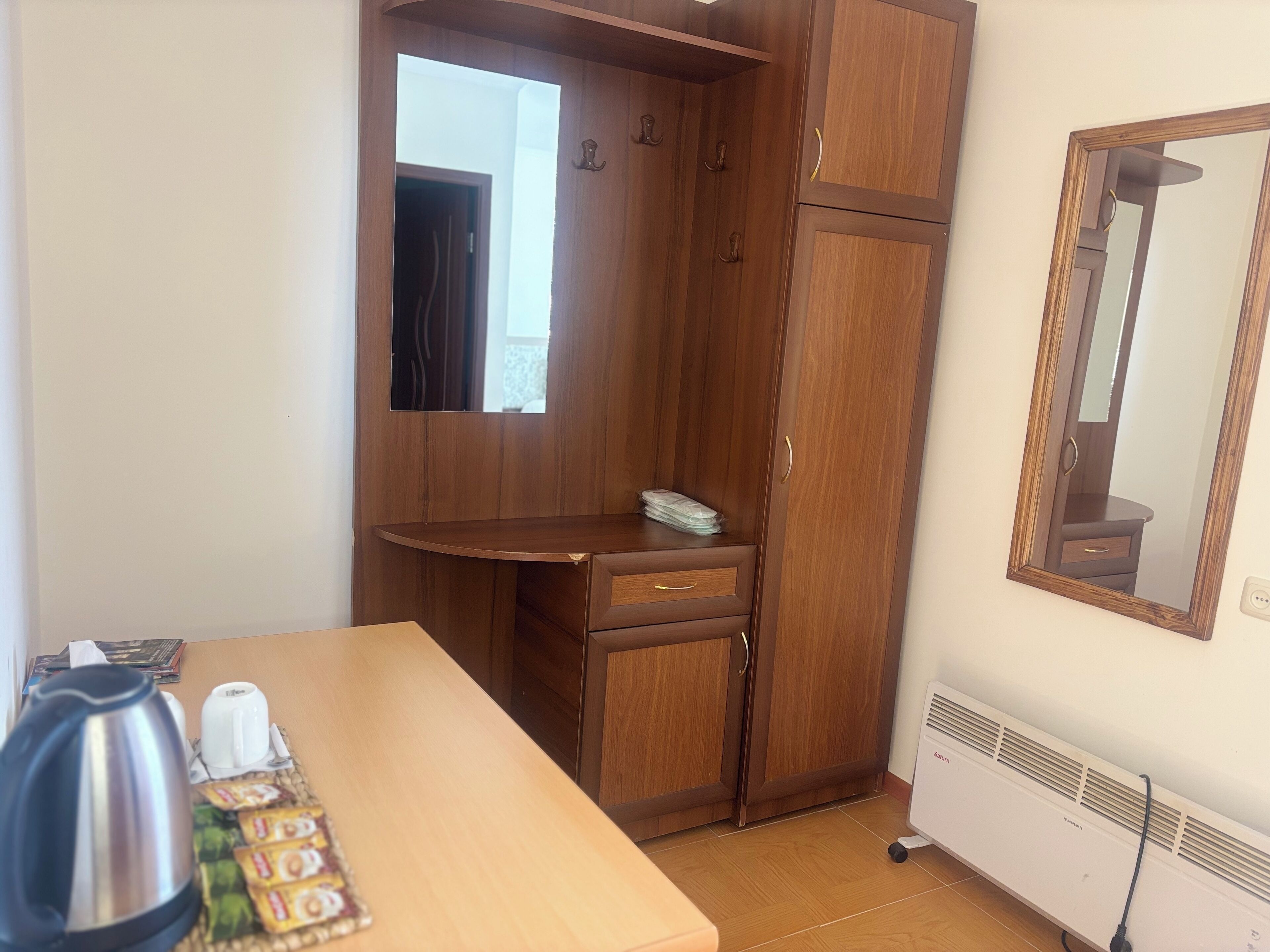 Een kamer met een houten garderobe, een spiegel, een tafel met waterkoker en theezakjes, en een radiator.