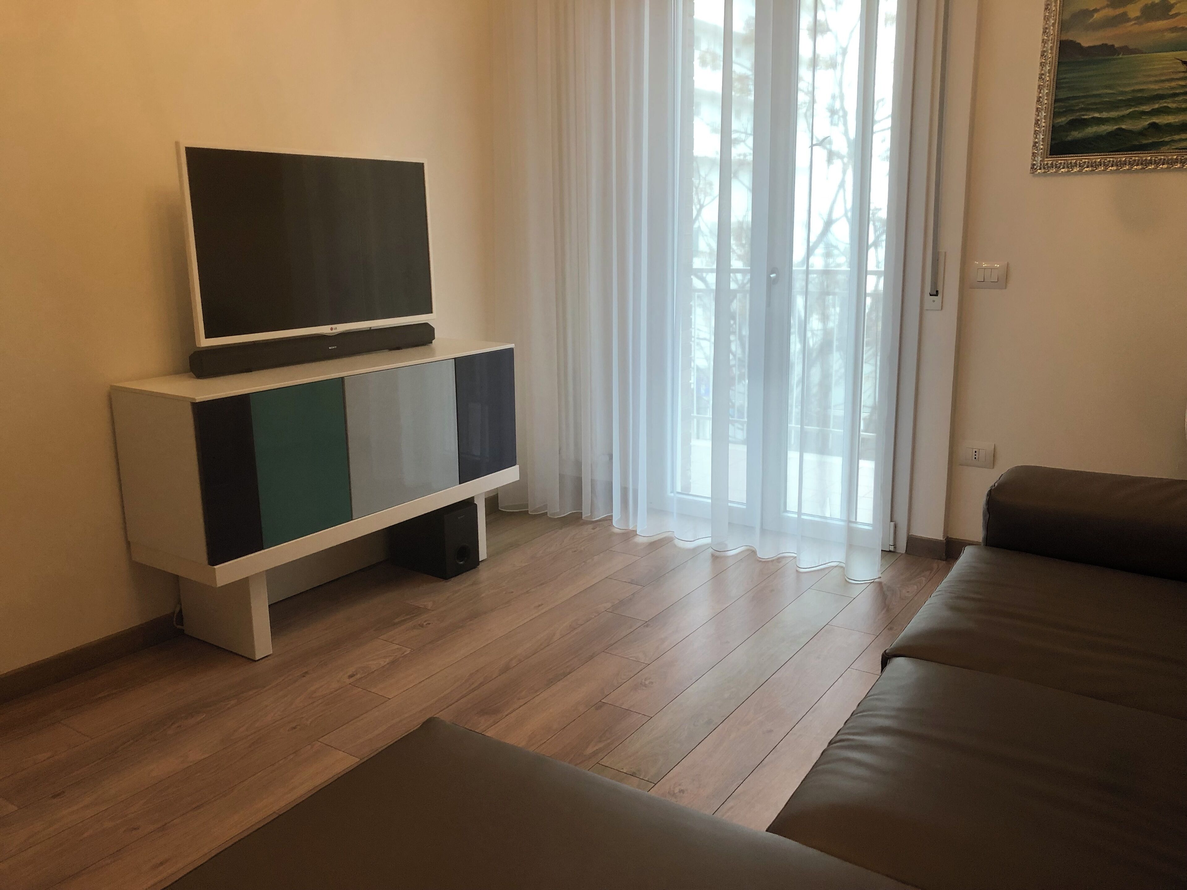 A living room with a flat-screen TV on a white cabinet, a brown sofa, and a large window with sheer curtains.