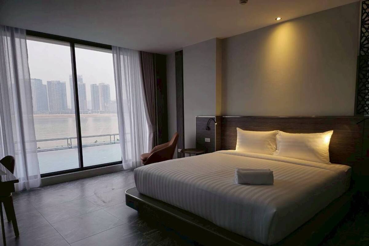 A hotel room with a large bed, a chair, a desk, and a view of a cityscape through large windows.