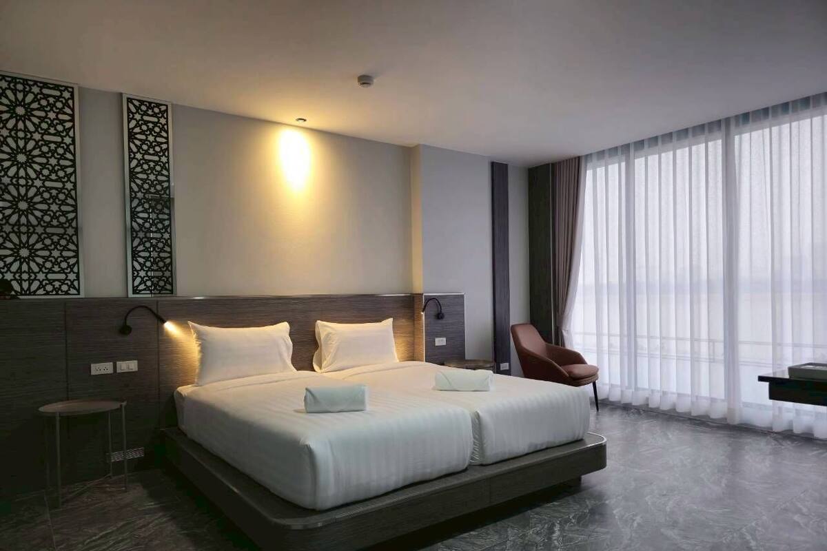 A modern hotel room with a large bed, a bedside table, a chair, and a view of the city through large windows.
