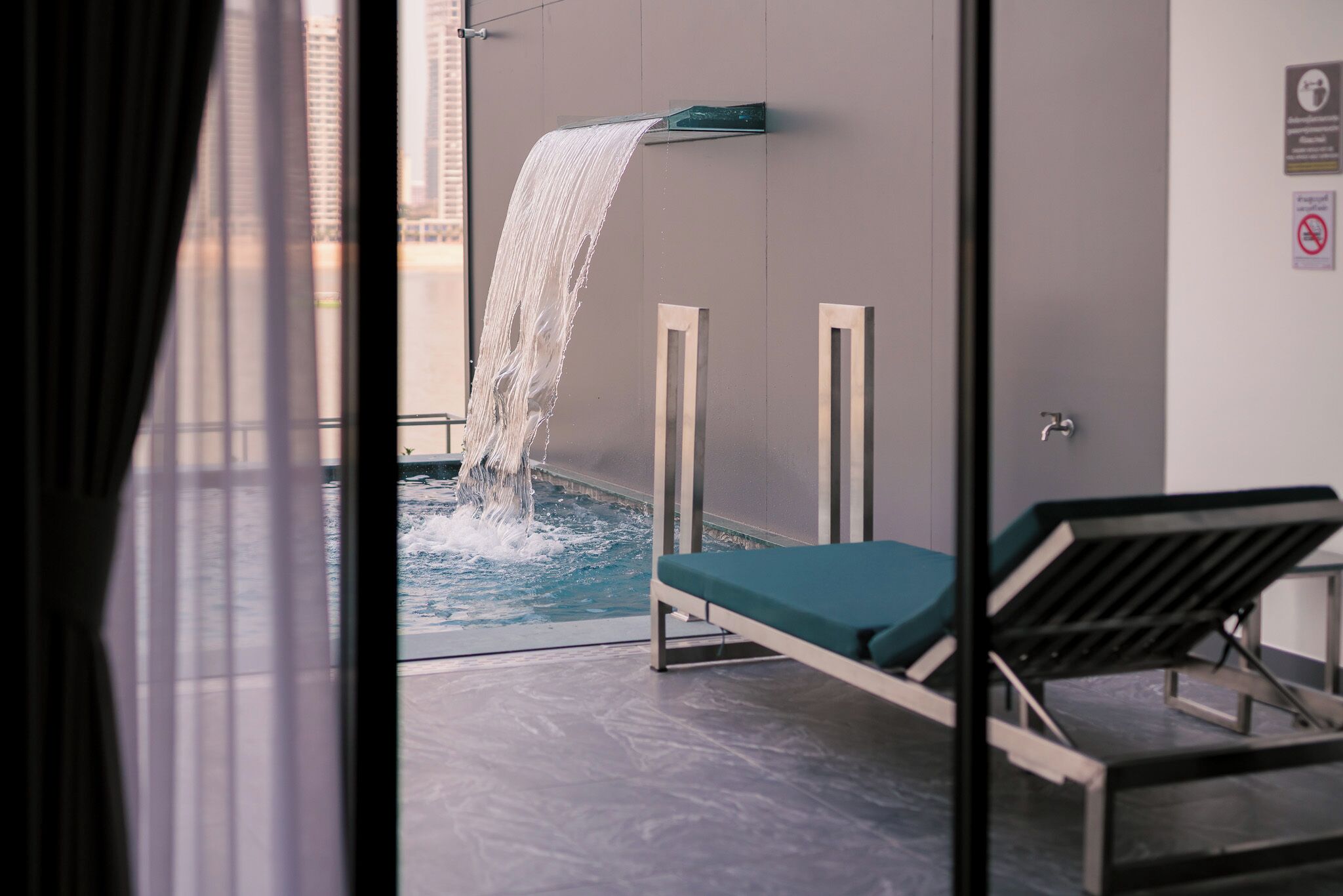 En moderne swimmingpool indendørs med et vandfald, en liggestol og udsigt til høje bygninger.
