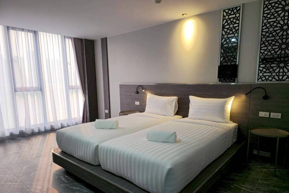 A double bed with white linens, a headboard, a bedside table, and a large window with sheer curtains.