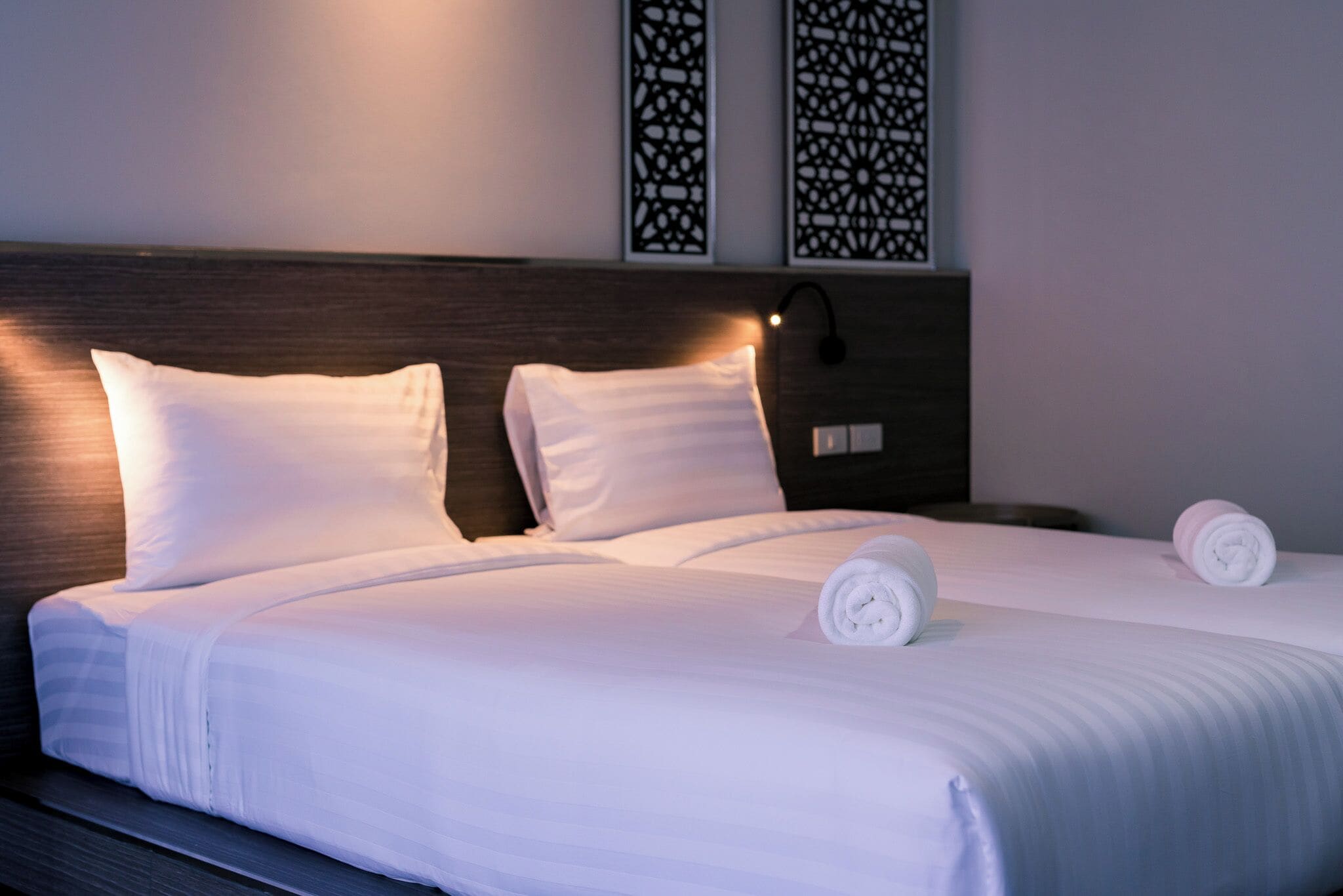 A neatly made bed with white linens, two pillows, and two rolled towels.